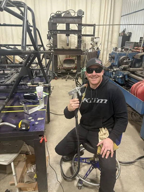 Scott Vogel seated in the fab shop holding a TIG torch, tube chassis on the fixture table, press and lathe behind